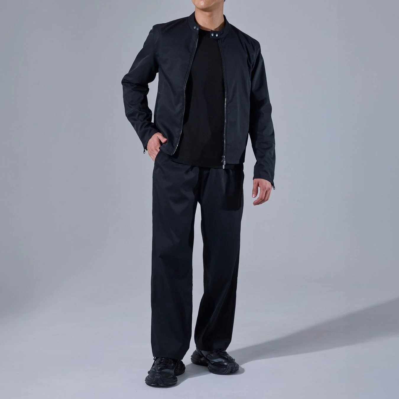 黒い襟なしジャケット、Tシャツ、ワイドパンツ、スニーカーを着用した男性の全身像